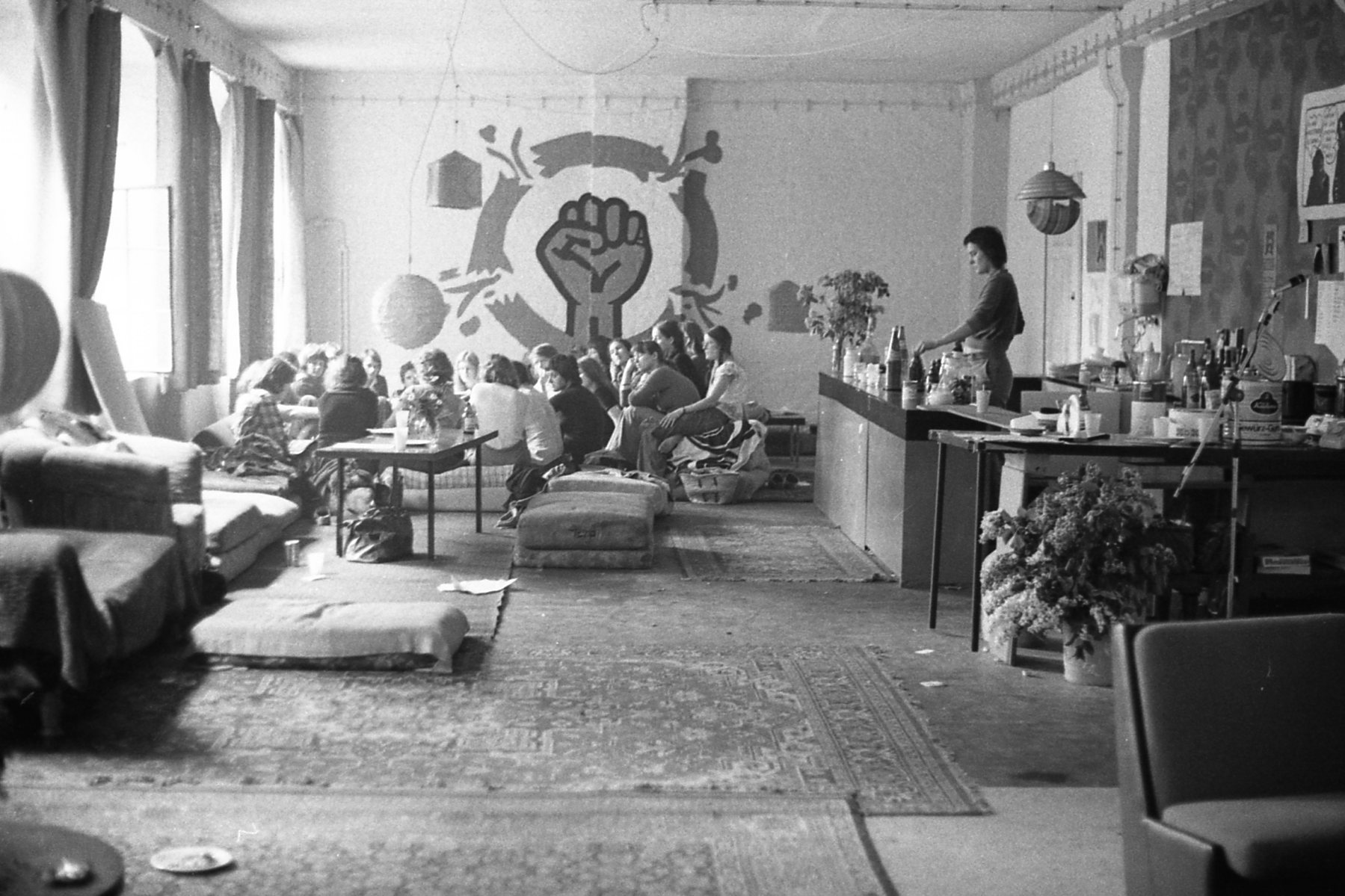 Schwarz-Weiß Fotografie des Gruppentreffen im LAZ, Kulmerstraße, Berlin-Schöneberg, 1975. Auf dem Foto ist eine Bar mit einer Person dahinter auf er rechten Bildseite zu sehen. Auf der linken Bildseite sitzt eine Gruppe von Personen zusammen. Auf der rückliegenden Wand ist eine übergroße, geballte Faus aufgemalt, die den Kreis sprengt, in dem sie sich befindet.