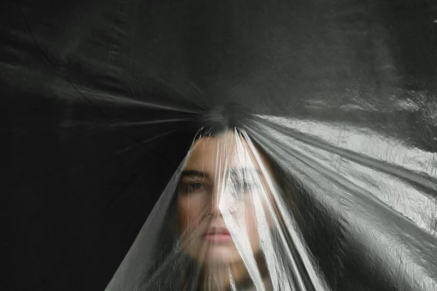 Ein weiblich gelesenens Gesicht ist unter einer Plastikfolie verschwommen zu sehen. Sie scheint wie vor äüßeren Einflüssen geschützt - verwart zu sein. 