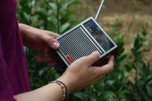 Das Bild zeigt eine Person in lila Tshirt, die ein altes Radio in Händen hält. Im Hintergrund ist eine Grünfläche zu sehen
