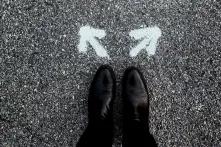 Foto von oben auf zwei Schuhe, die auf einer Straße stehen, auf dem Boden vor den Schuhspitzen sind jeweils zwei weiße Pfeile nach rechts und links oben gemalt.