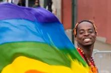 Eine Schwarze Person lächelt in die Kamera und schwingt dabei eine große Regenbogen Flagge.