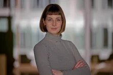 Portraitfoto Tini Leonhardt - eine weiblich gelesene Person mit kinnlangem hellen Haar steht mit verschränkten Armen und zugewandt zur Kamera vor einem eher verschwommenen Hintergrund.