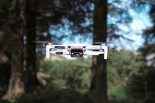 eine weiße Drohne mit Kamera fliegt direkt auf die das Bild betrachtende Person zu, im Hintergrund stehen Bäume. Der Winkel lässt darauf schliessen dass die Drohne die betrachtende Person auf Augenhöhe konfrontiert.