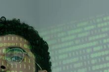 Ein Auge eines männlich gelesenen Gesichtes blickt uns durch eine Brille hinweg an, auf das Gesicht und den neutralen grauen Hintergrund sind grüne Zahlen projiziert, die an einen Computer-Code erinnern