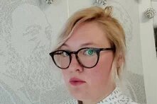 eine weiße weiblich gelesene Person mit Brille und mit platinblonden Haaren blickt im 3/4-Profil ernst in die Kamera. Sie ist etwa mitte dreißig. Hinter ihr hängt ein Plakat mit Karl Marx. Das Bild ist sehr hell, Weiß und Beigetöne dominieren.