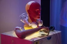 Humanoide Bauteile -ein Arm, eine Büste - jeweils aus Kunststoff, ähnlich einer Schaufensterpuppe sind auf einem Sockel verbaut. Der Arm versucht mit zwei Fingern ein Kärtchen zu greifen , um den Hals ist ein Kopfhörer gelegt. Durchsichtige Schläuche, Ähnlich wie Katheter im Krankenhaus verbinden Arm und Kopf. Das Arragement ist ein Kunstwerk einer Ausstellung.