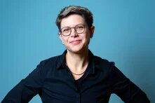 Vor blauem Grund steht, die Hände in die Hüften gestützt, schwarz gekleidet eine schlanke Person in ihren frühen 40ern. Die Haare sind kurz, der Blick durch eine Brille lächelt Betrachtende direkt an. 