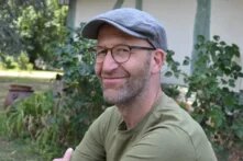 Ein weißer Mann mittleren Alters sitzt in einem sommergrünen Garten. Er trägt Brille und Schiebermütze und lächelt im Halbprofil in die Kamera.