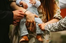 Ein Baby wird von zwei weißen weiblichen Händen gehalten. Eine weitere Hand nähert sich fürsorglich dem Bein des Kindes. Ein weiteres Händepaar ist ganz in seiner Nähe. Die Szene wirkt wohlig, kümmernd, dynamisch.