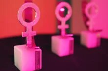 Drei Pokale des Anne-Klein-Fraunpreises (das Weiblichkeitssymbol auf einem kleinen Marmor-Podest) stehen in einer reihe in rosa Licht getaucht.