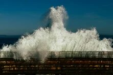 Eine tosende Welle bricht über eine Barriere hinein. Sie schäumt weiss und kommt frontal auf uns zu. 