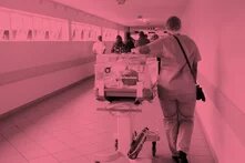 Durch einen Krankenhauskorridor wird ein Kleinkindbett von einer Person in OP-Kittel geschoben. Alles ist durch einen altrosa Filter gefärbt.