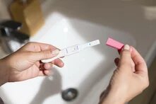 Ein Schwangerschaftstest ist positiv ausgefallen. Zwei weisse weibliche Hände halten ihn über einem sonnenbeschienen Waschbecken. Es wirkt wie eine Point-of-View-Aufnahme.