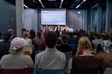 Foto aus Publikumssicht ganz hinten: Vorme auf der Bühne sitzen 5 Personen mit einer Leinwand, wo Titel der Veranstaltung und Namen der Panelist*innen stehen. Der Raum ist hell beleuchtet und grau.