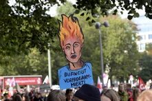 Ein Plakat auf einer Demo steht im Mittelpunkt: die Haare einer erschöpften Menschlichen Figure "stehen in Flammen", darunter steht: Burnout vorprogrammiert.