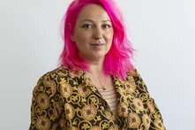 Eine junge weibliche Person in gelb gemustertem Blazer und pinken Haaren blickt freundlich und selbstbewusst in die Kamera.