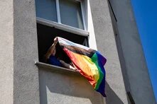 Aus einem Eckhausfenster schüttelt ein:e junge:r Aktivist:in eine progressive Queerflagge in den blauen Sommerhimmel, wie Bettzeug zum Lüften. Das Bild enstand beim CSD 2025 in Eberswalde.