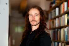 Ein Mann in seinen Vierzigern blickt uns im Halbprofil an. Er hat langes gewelltes kastanienbraunes Haar, hellblaue Augen und markante Wangenknochen. Hinter ihm erkennt man eine voll bestückte Bücherwand.