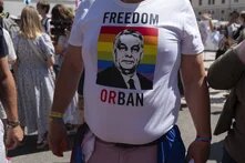 Eine Person steht mit geschwellter Brust und trägt ein weißes Tshirt. Auf das Shirt ist eine Prideflagge mit dem Gesicht von Viktor Orban gedruckt. Über dem Druck steht "FREEDOM" und unter dem Druck "OR BAN"