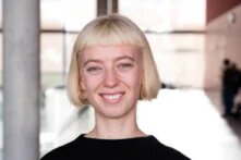 Eine junge weisse Person mit platinblondem Bob lächelt in die Kamera. Sie ist schlank, trägt schwarz und ist zwischen 25 und 35 Jahre alt.