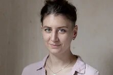 Pascale Müller, eine Person mit dunklen hochgesteckten Haaren und hellem Hemd steht lächelnd vor einer beigen Wand.