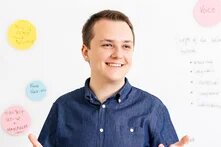 Person in blauem Hemd steht lächelnd und mit Händen gestikulierend vor einer Wand mit Moderationskarten