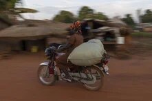 Eine Frau auf einem Motorrad, auf dem sie einen Sack transportiert, Ortsansicht und Details in Nzara im Suedsudan.