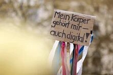 Ein Schild mit der Aufschrift: "Mein Körper gehört mir - auch digital!" wird hochgehalten. An dem Schild hängen bunte Streifen hinunter.