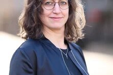 zu sehen ist die Autorin Johanna Wies im Portrait, schulterlange braune Haare, Brille, blaue Jacke über blauem Shirt mit kurzer Kette;