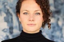 Eine weiblich gelesene Person sieht mit blaugrauen Augen direkt die betrachtende Person an. Sie hat braune gelockte Haare, die sie hochgesteckt trägt. Der Gesichtsausdruck ist ruhig und selbstsicher.