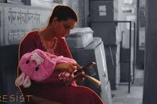 Photo: A woman in traditional clothing sits on a bench, holding a baby wrapped in a pink blanket. She looks at her phone while holding an open book. The word "RESIST" is written in the image.