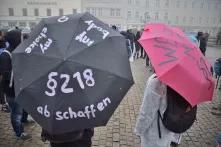 Zwei Personen mit Regenschirmen stehen mit dem Rücken zur Kamera. Auf einem Regenschrim steht §218 abschaffen.