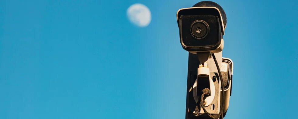 Blauer Himmer, Mond in Tiefenunschärfe, davor eine Kamera auf einem Mast staffiert die die Betrachtenden fokussiert