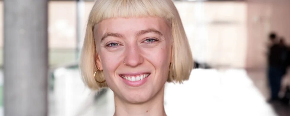 Eine junge weisse Person mit platinblondem Bob lächelt in die Kamera. Sie ist schlank, trägt schwarz und ist zwischen 25 und 35 Jahre alt.