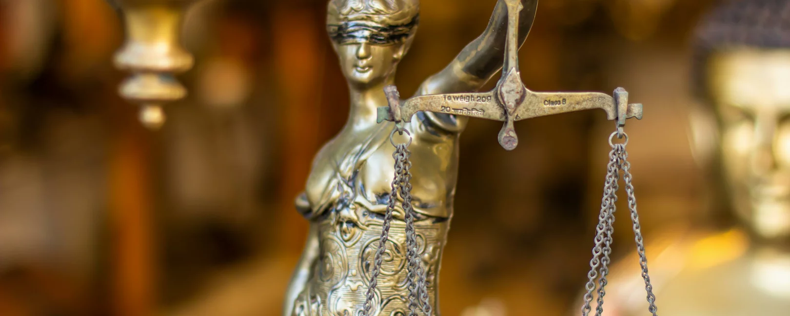 Es wird eine Justicia-Figur in goldener Farbe im Anschnitt dargstellt. Der Hintergrund ist verschwommen.