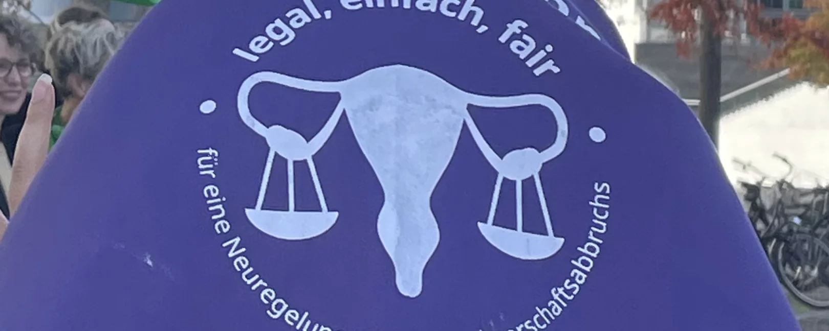 Eine Person steht mit dem Rücken zu uns und trägt ein lilanes dreieckiges Kopftuch mit der Aufschrift 