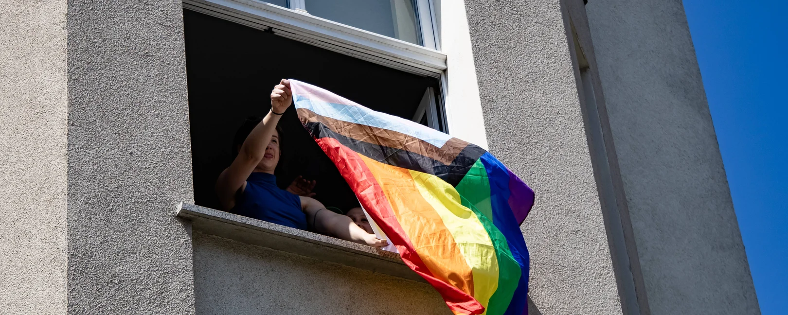 Aus einem Eckhausfenster schüttelt ein:e junge:r Aktivist:in eine progressive Queerflagge in den blauen Sommerhimmel, wie Bettzeug zum Lüften. Das Bild enstand beim CSD 2025 in Eberswalde.