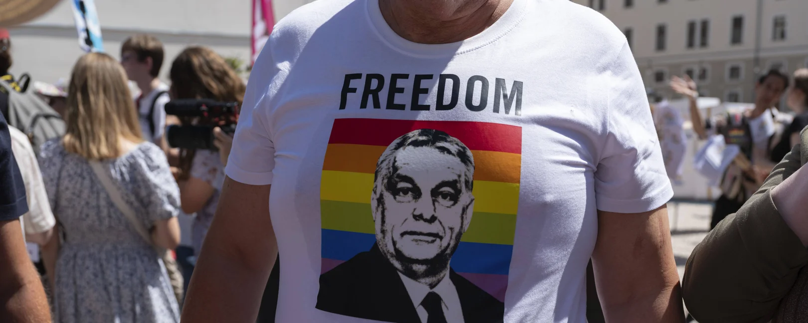 Person trägt ein weißes Shirt mit folgendem Print: Regenbogenflagge und ein Portrait von Viktor Orban. Über dem Print steht in großen schwarzen Buchstaben 