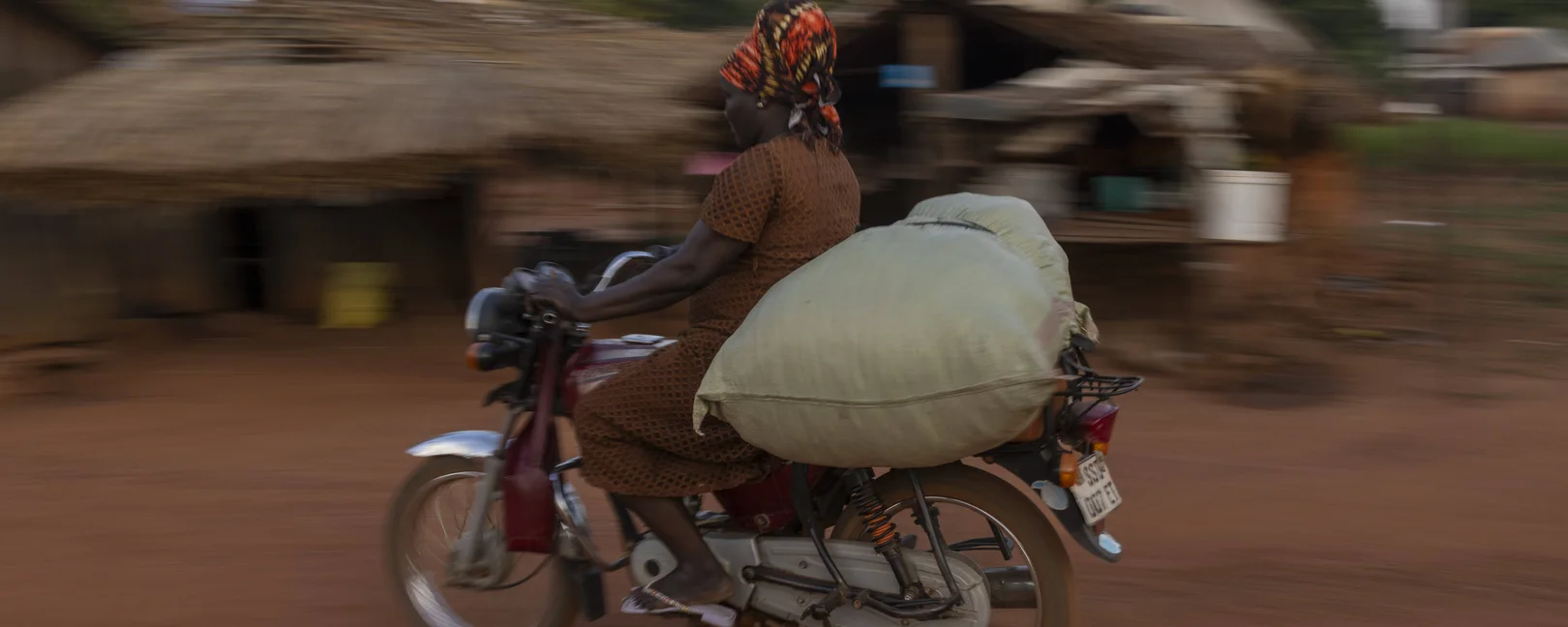Eine Frau auf einem Motorrad, auf dem sie einen Sack transportiert, Ortsansicht und Details in Nzara im Suedsudan.