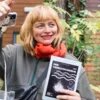 Eine weiße, blonde Frau um die 40 gestikuliert ausschweifend während sie etwas einem Publikum ausserhalb der Bildes zu erklären scheint. Sie sieht freundlich und etwas verschmitzt aus. Ihre Haare sind zu einem Bob mit geradem Pony geschnitten. Ihre Augen sind grünblau. Sie trägt roten Lippenstift, eine Bomberjacke und einen orangen Schal. Alisa Tretau