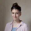Pascale Müller, eine Person mit dunklen hochgesteckten Haaren und hellem Hemd steht lächelnd vor einer beigen Wand.