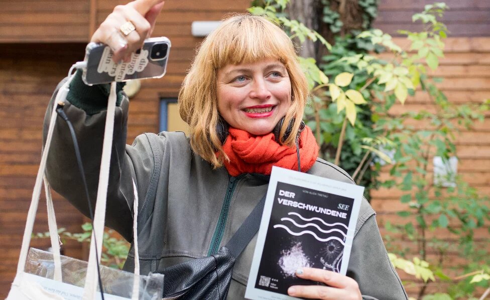 Eine weiße, blonde Frau um die 40 gestikuliert ausschweifend während sie etwas einem Publikum ausserhalb der Bildes zu erklären scheint. Sie sieht freundlich und etwas verschmitzt aus. Ihre Haare sind zu einem Bob mit geradem Pony geschnitten. Ihre Augen sind grünblau. Sie trägt roten Lippenstift, eine Bomberjacke und einen orangen Schal. Alisa Tretau