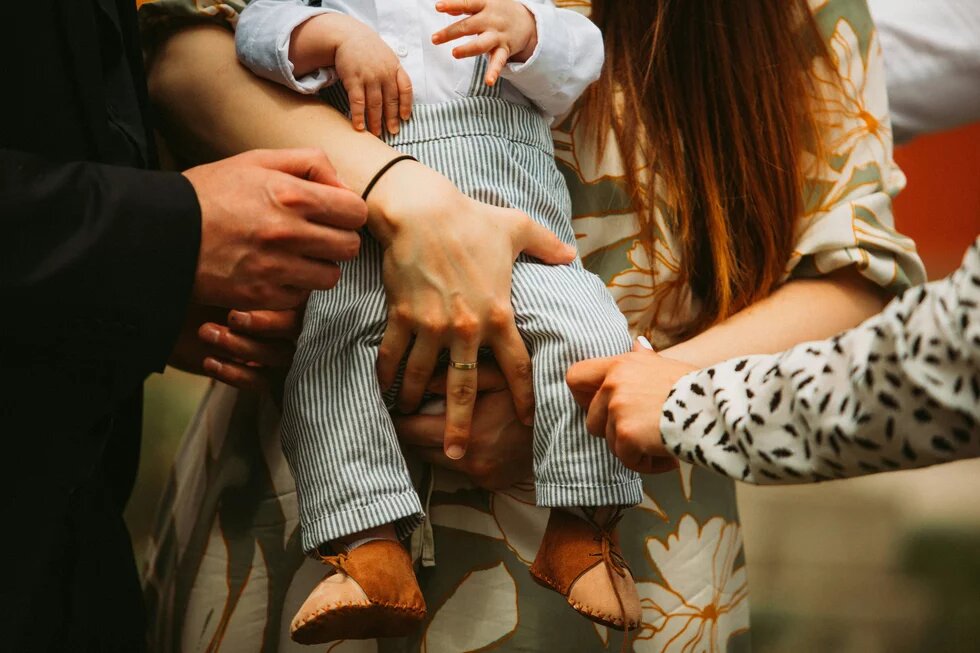 Ein Baby wird von zwei weißen weiblichen Händen gehalten. Eine weitere Hand nähert sich fürsorglich dem Bein des Kindes. Ein weiteres Händepaar ist ganz in seiner Nähe. Die Szene wirkt wohlig, kümmernd, dynamisch.