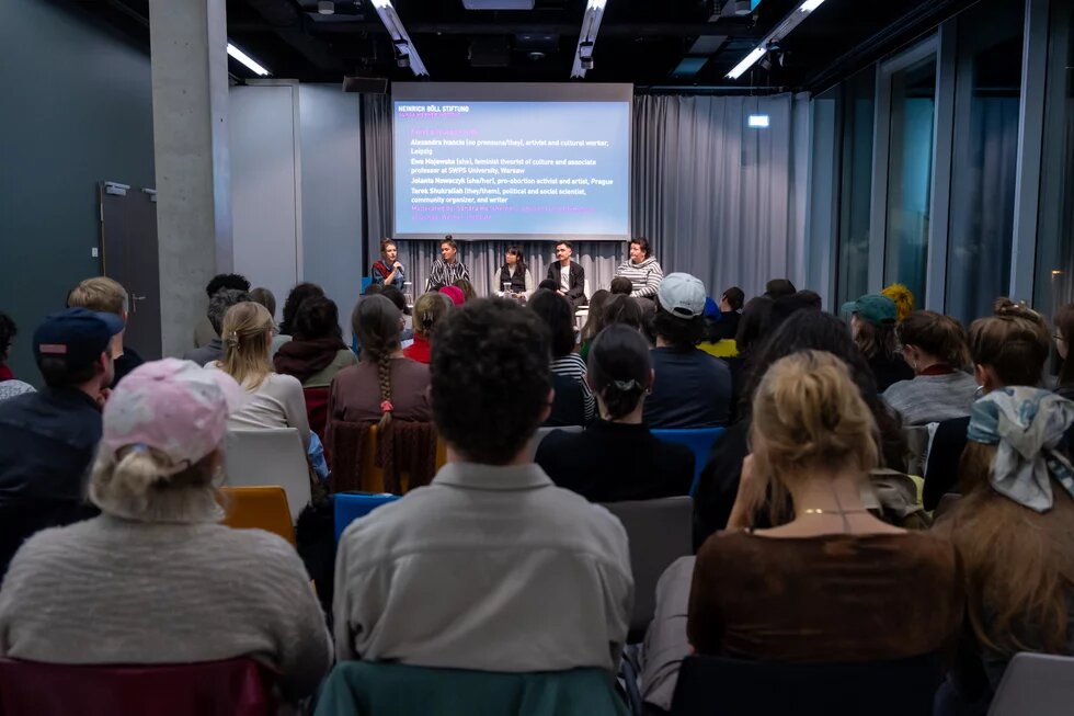 Foto aus Publikumssicht ganz hinten: Vorme auf der Bühne sitzen 5 Personen mit einer Leinwand, wo Titel der Veranstaltung und Namen der Panelist*innen stehen. Der Raum ist hell beleuchtet und grau.