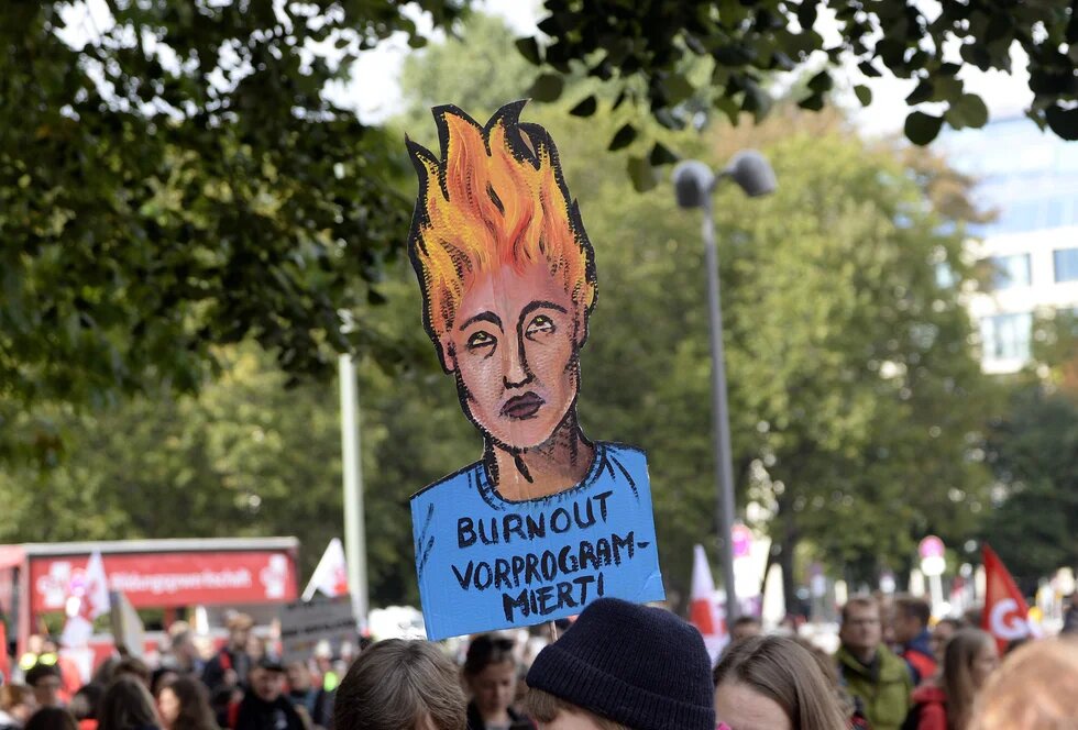 Ein Plakat auf einer Demo steht im Mittelpunkt: die Haare einer erschöpften Menschlichen Figure "stehen in Flammen", darunter steht: Burnout vorprogrammiert.