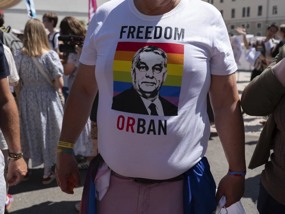 Eine Person steht mit geschwellter Brust und trägt ein weißes Tshirt. Auf das Shirt ist eine Prideflagge mit dem Gesicht von Viktor Orban gedruckt. Über dem Druck steht "FREEDOM" und unter dem Druck "OR BAN"