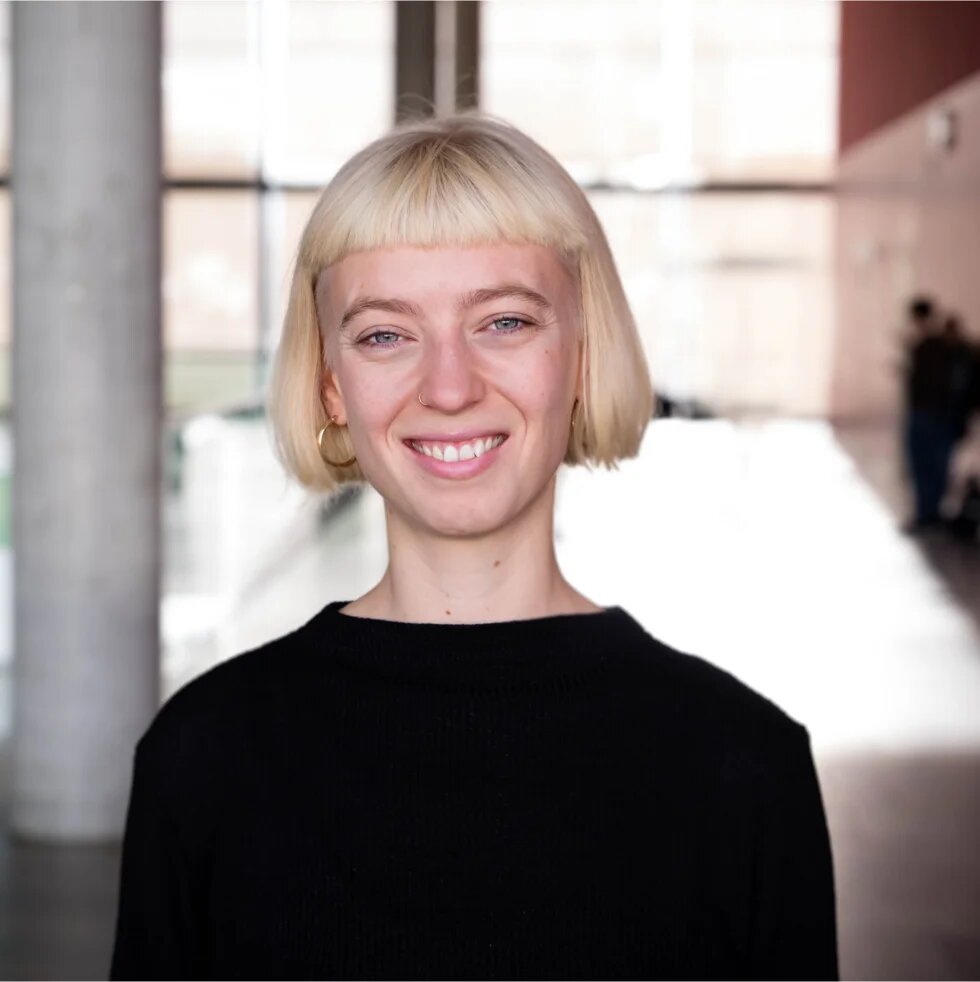 Eine junge weisse Person mit platinblondem Bob lächelt in die Kamera. Sie ist schlank, trägt schwarz und ist zwischen 25 und 35 Jahre alt.