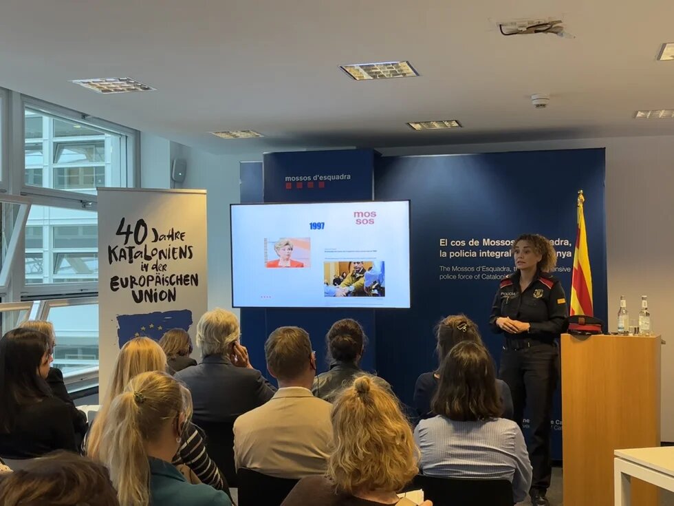 Korporal Eva Martín, Polizeibeamtin der Mossos d'Esquadra, hält eine Präsentation zum Gewaltschutzsystem. Im Hintergrund steht ein Aufsteller mit der Aufschrift "40 Jahre Kataloniens in der Europäischen Union.". 