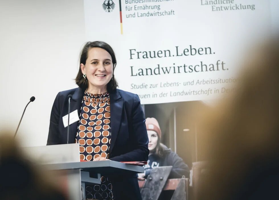Person steht an einem Rednerpult. Dahinter ist eine Präsentation mit der Aufschrift "Frauen, Leben, Landwirtschaft" dargestellt.