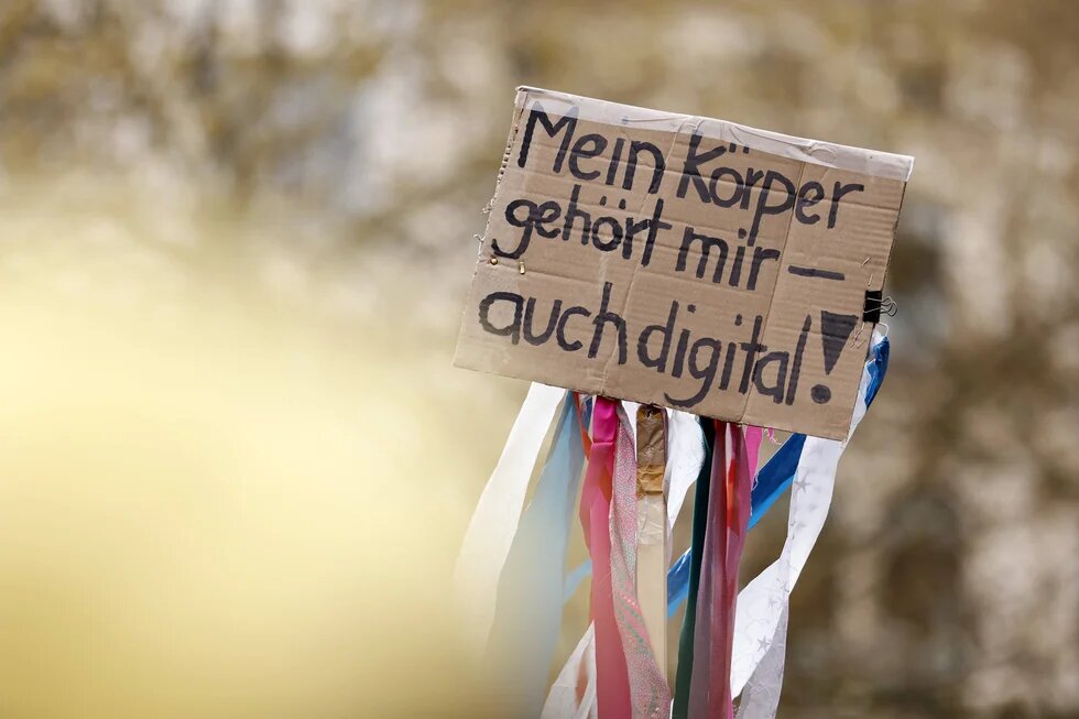 Ein Schild mit der Aufschrift: "Mein Körper gehört mir - auch digital!" wird hochgehalten. An dem Schild hängen bunte Streifen hinunter.